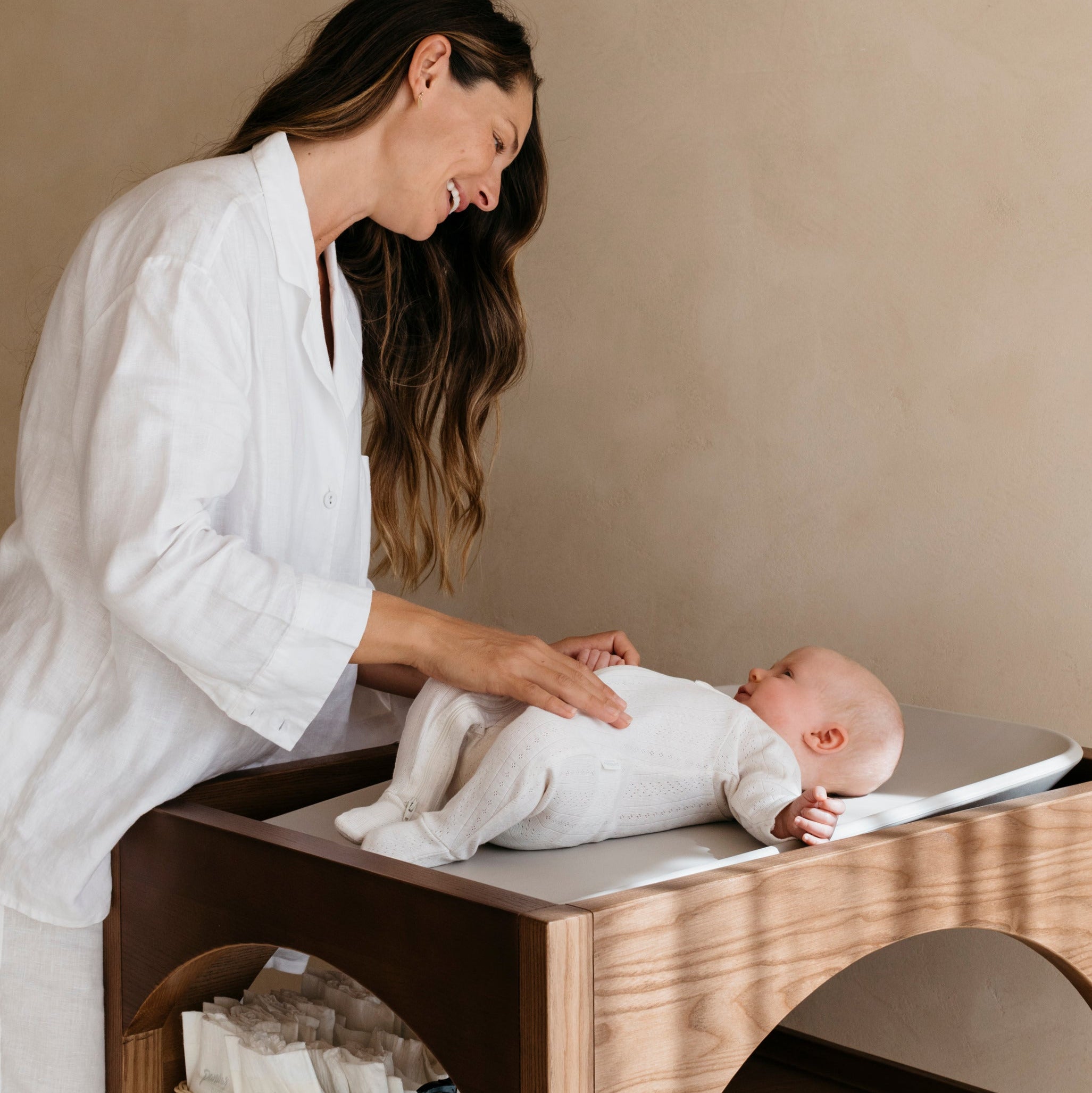 Woman changing a baby on a wooden changing table against a beige wall.