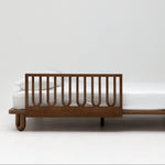 A modern wooden kids bed frame with a side rail, shown with a white mattress and pillows against a clean white studio background.