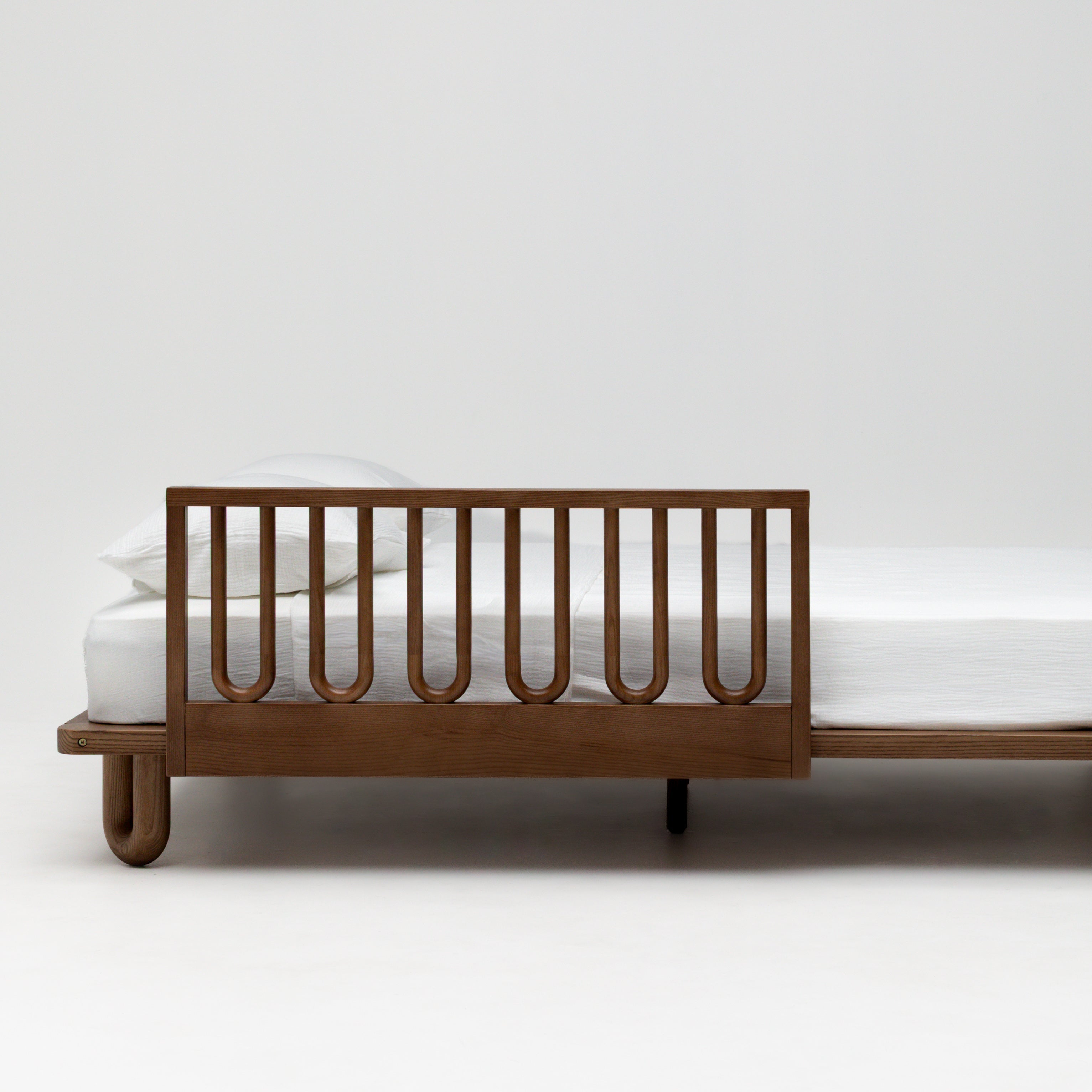 A modern wooden kids bed frame with a side rail, shown with a white mattress and pillows against a clean white studio background.