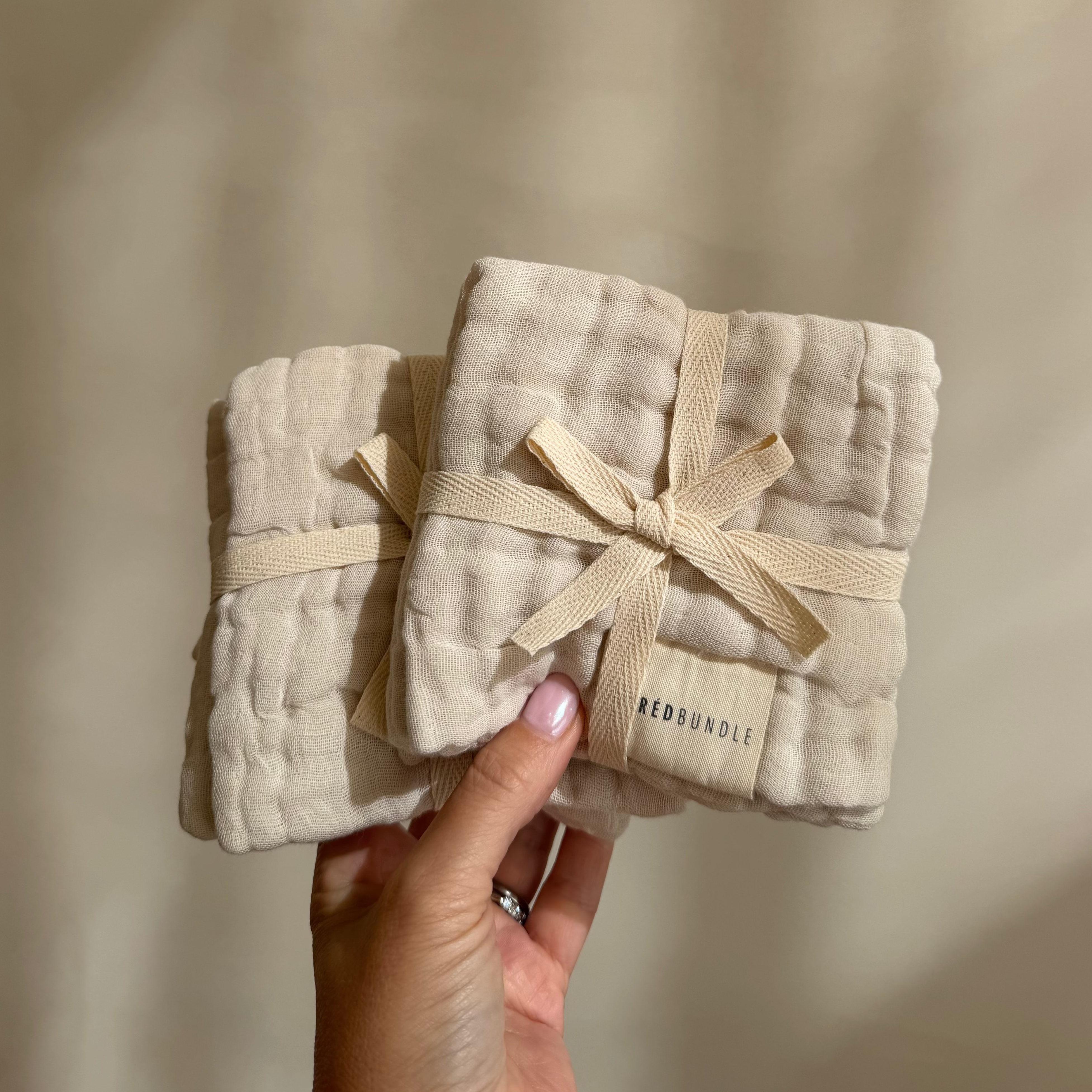 Two beige fabric wash cloth tied with ribbons held by a hand against a neutral background