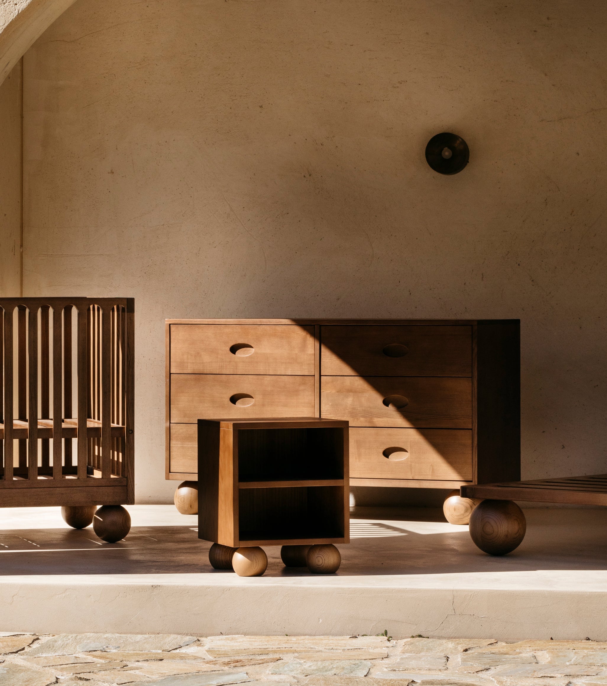 Wooden walnut nursery furniture pieces including a cot, buffet dresser, side table and kids single bed against a concrete wall.