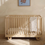 A light natural wood crib with curved legs and arched frames, shown in a softly lit nursery with a cream wall, framed wall art, and a patterned baby blanket draped over the side.