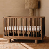 A wooden crib with vertical cot slats and cylinder legs, shown with a white cot mattress in a warm neutral nursery room with a textured wall and floor lamp.