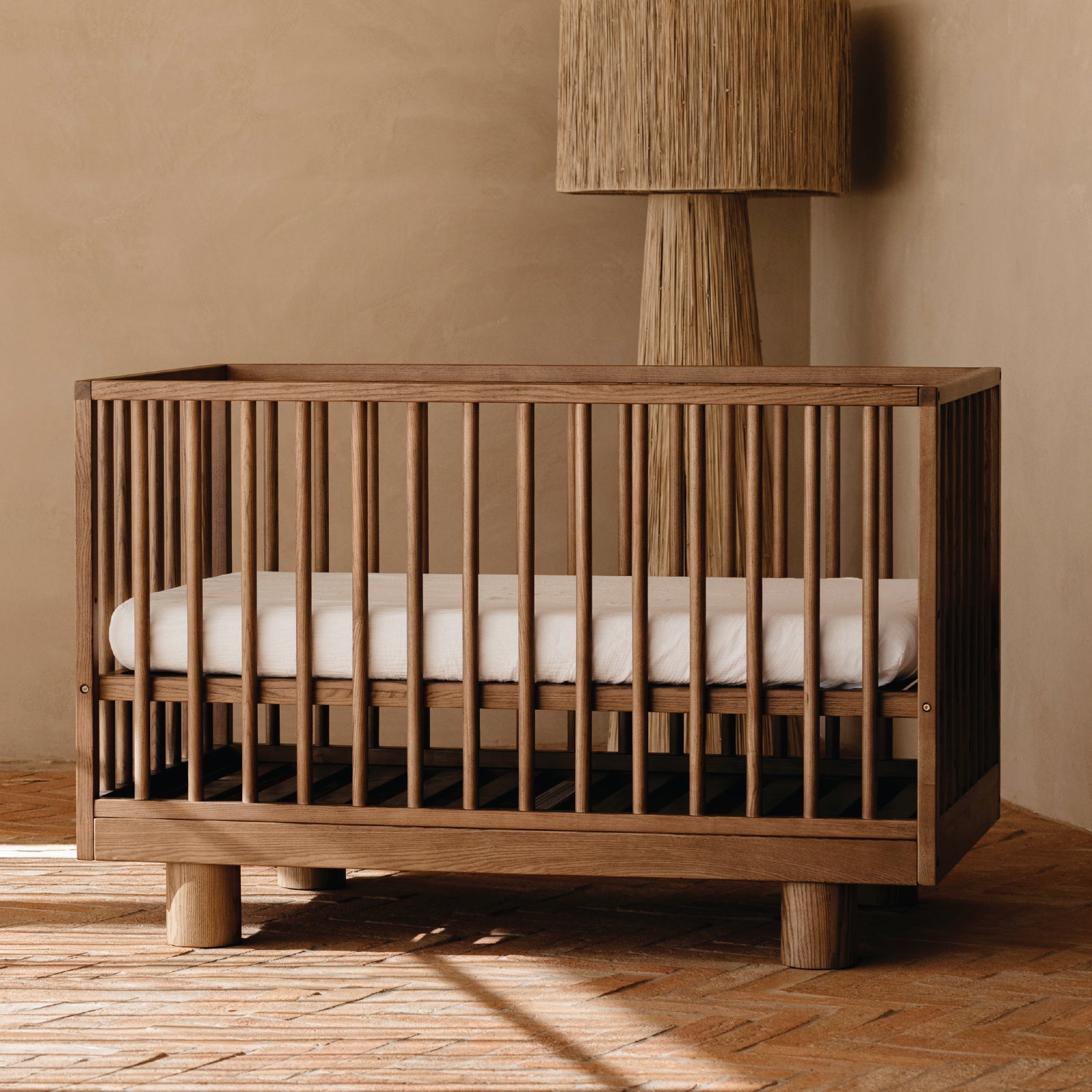 A wooden crib with vertical cot slats and cylinder legs, shown with a white cot mattress in a warm neutral nursery room with a textured wall and floor lamp.