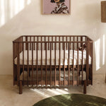A walnut wooden baby crib with vertical cot slats and cot mattress, styled in a warm, sunlit nursery with a cream-colored wall, a framed art print, and a green rug in the foreground.