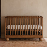 A wooden walnut crib with vertical slats and round ball feet, shown with a white cot mattress in a warm, rustic nursery room with a textured beige wall and brick floor.