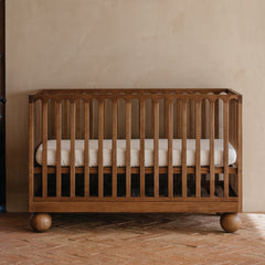 A wooden walnut crib with vertical slats and round ball feet, shown with a white cot mattress in a warm, rustic nursery room with a textured beige wall and brick floor.