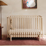 A light natural wood baby crib with vertical cot slats, styled in a nursery setting on a dusty pink rug.