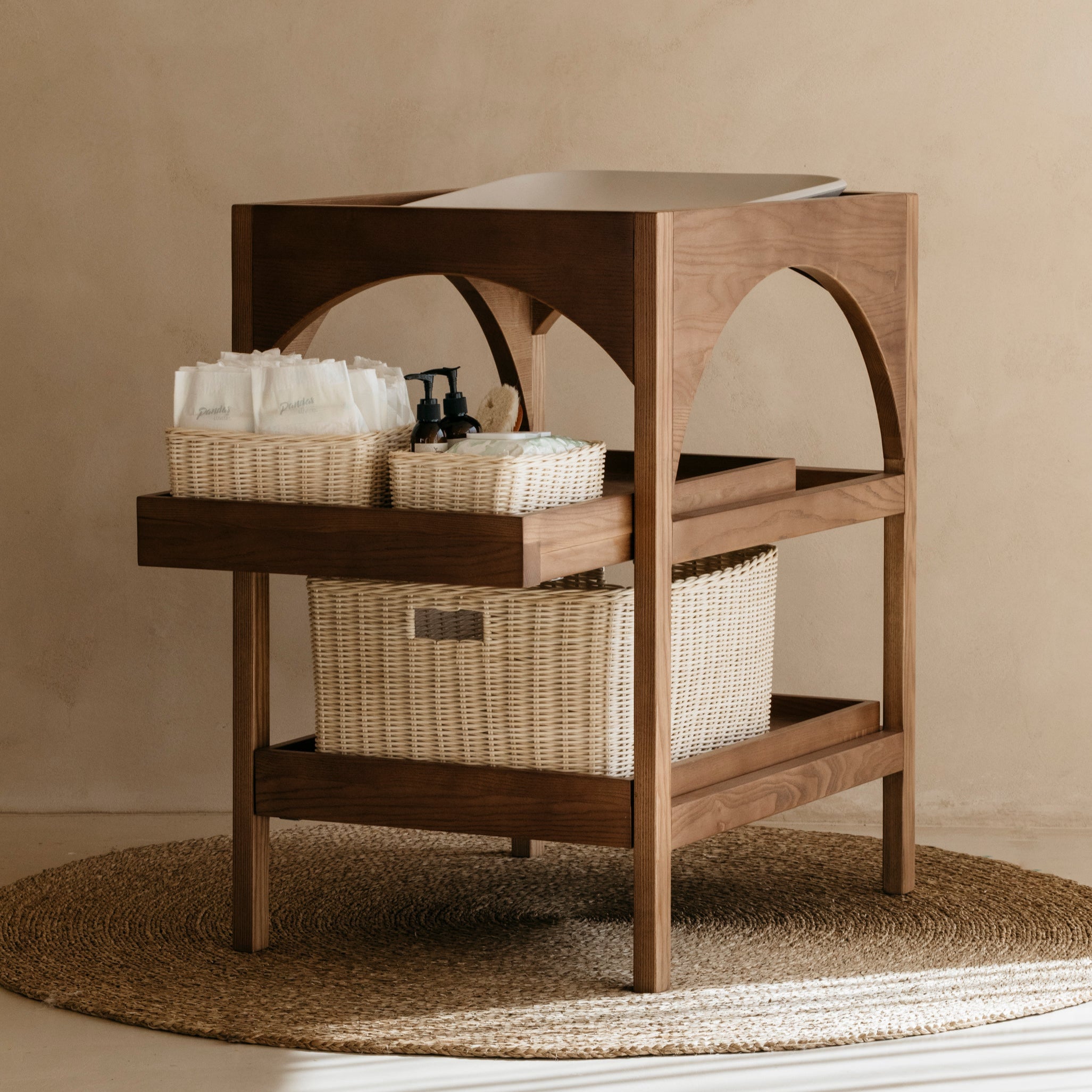 Walnut baby changing table with rattan baskets holding nappies and nursery essentials with change mat on top against a beige wall