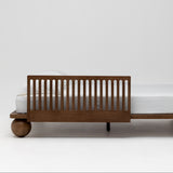 A mid-century modern wooden kids bed frame with a slatted side rail and spherical ball feet, shown in profile with a white mattress on a clean white studio background.